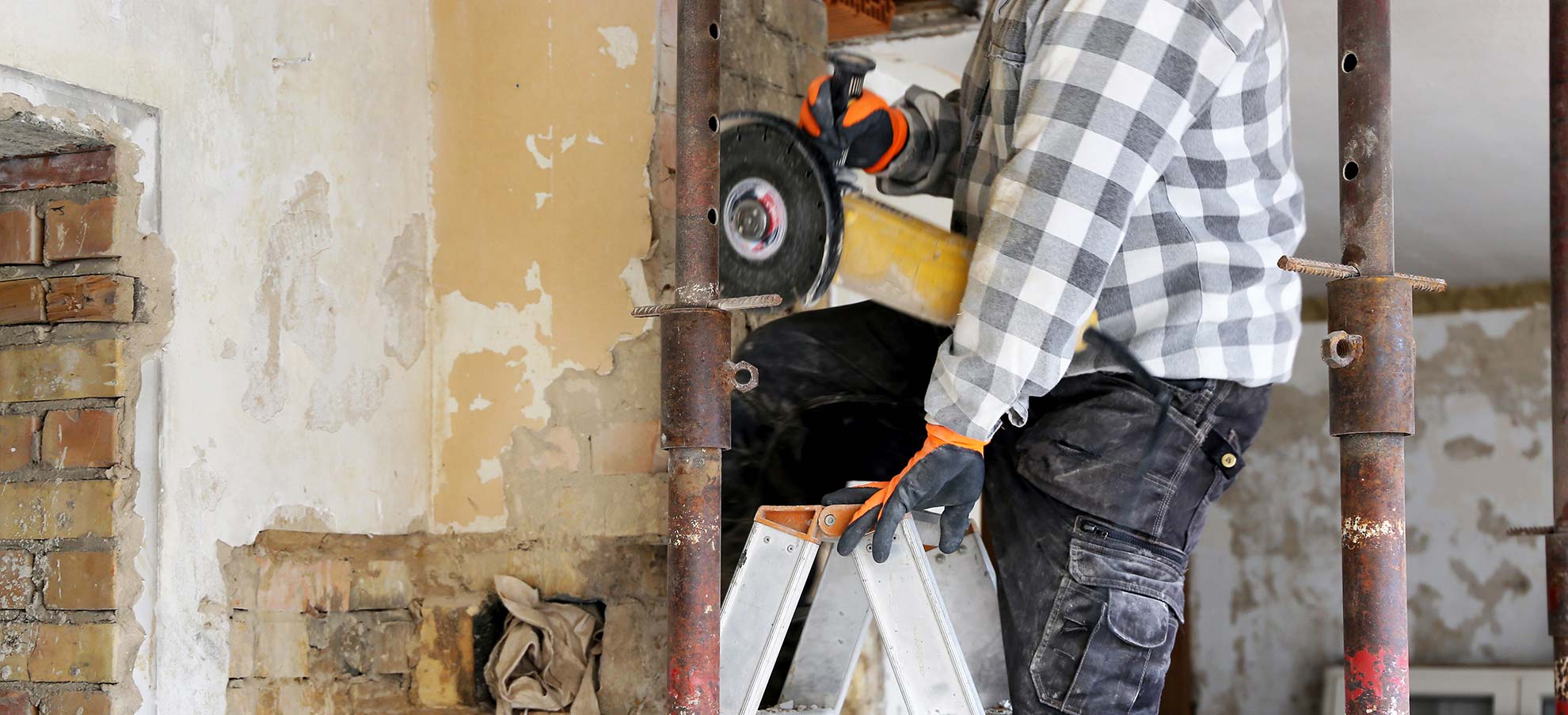 Ein Handwerker, von dem nur der Unterkörper sichtbar ist, steht auf einer Leiter in einem entkernten Gebäude mit einem Flex Werkzeug in der Hand.