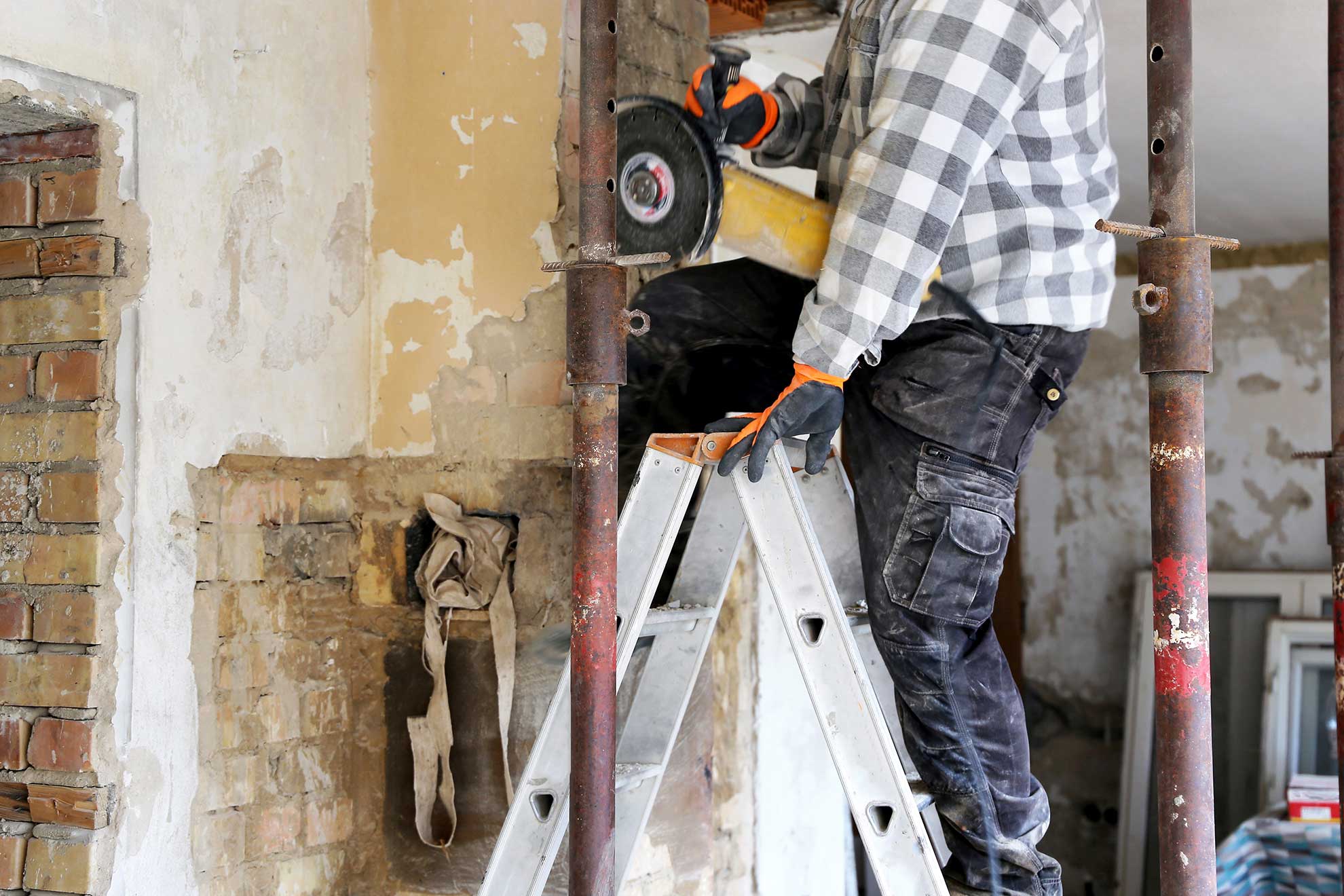 Ein Handwerker, von dem nur der Unterkörper sichtbar ist, steht auf einer Leiter in einem entkernten Gebäude mit einem Flex Werkzeug in der Hand.