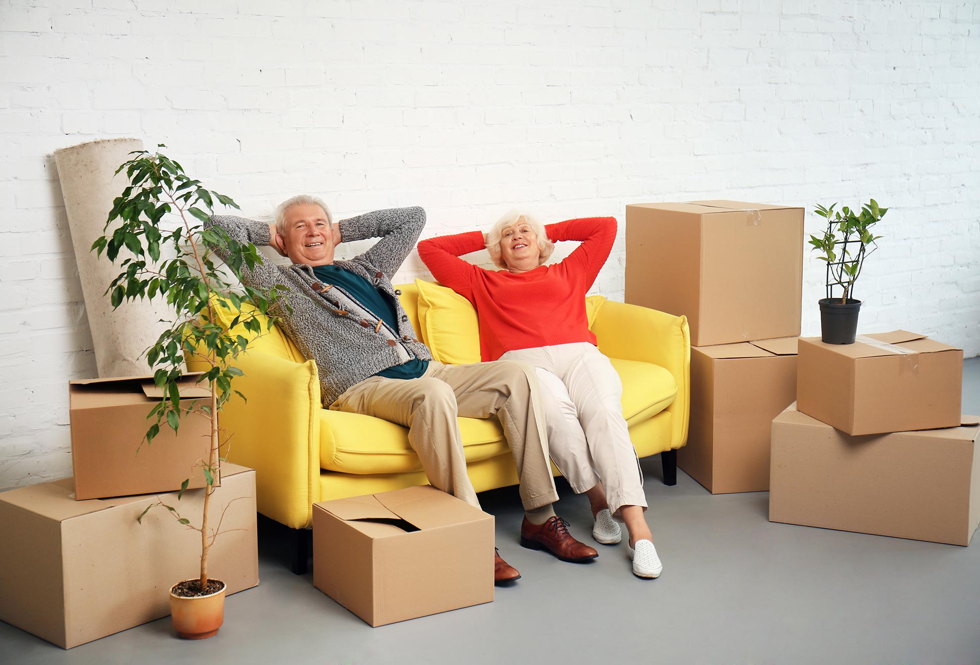Ein älteres Ehepaar sitzt fröhlich zurückgelehnt auf einer gelben Couch. Links und rechts befinden sich Umzugskartons und eine Pflanze.